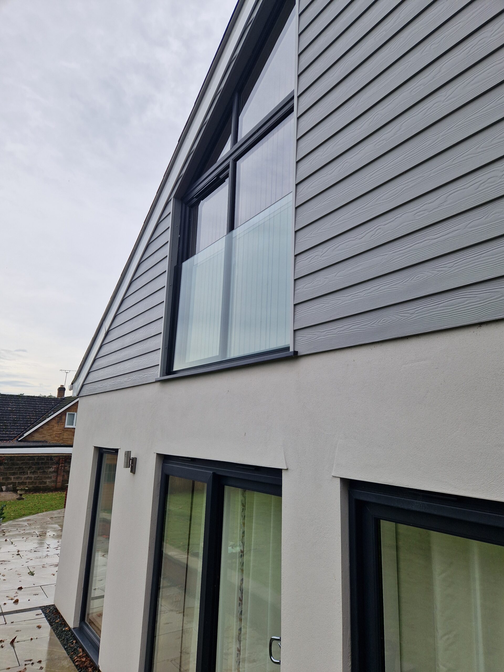 A Juliet balcony with a minimalist glass design, installed against a dark grey rendered wall with horizontal wood slats and a textured concrete base.